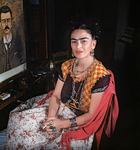 Photo of Frida Kahlo sitting next to a painting, adorned in traditional Mexican jewelry and clothing